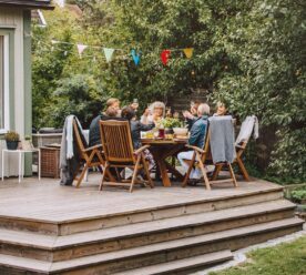 En folk mennesker sidder ved et spisebord terrasse og skåler glade med flag og lyskæder i træerne i baggrunden