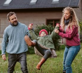 Mand og gravid kvinde løfter mindre pige med hue og gummistøvler op i armene i have foran et hus